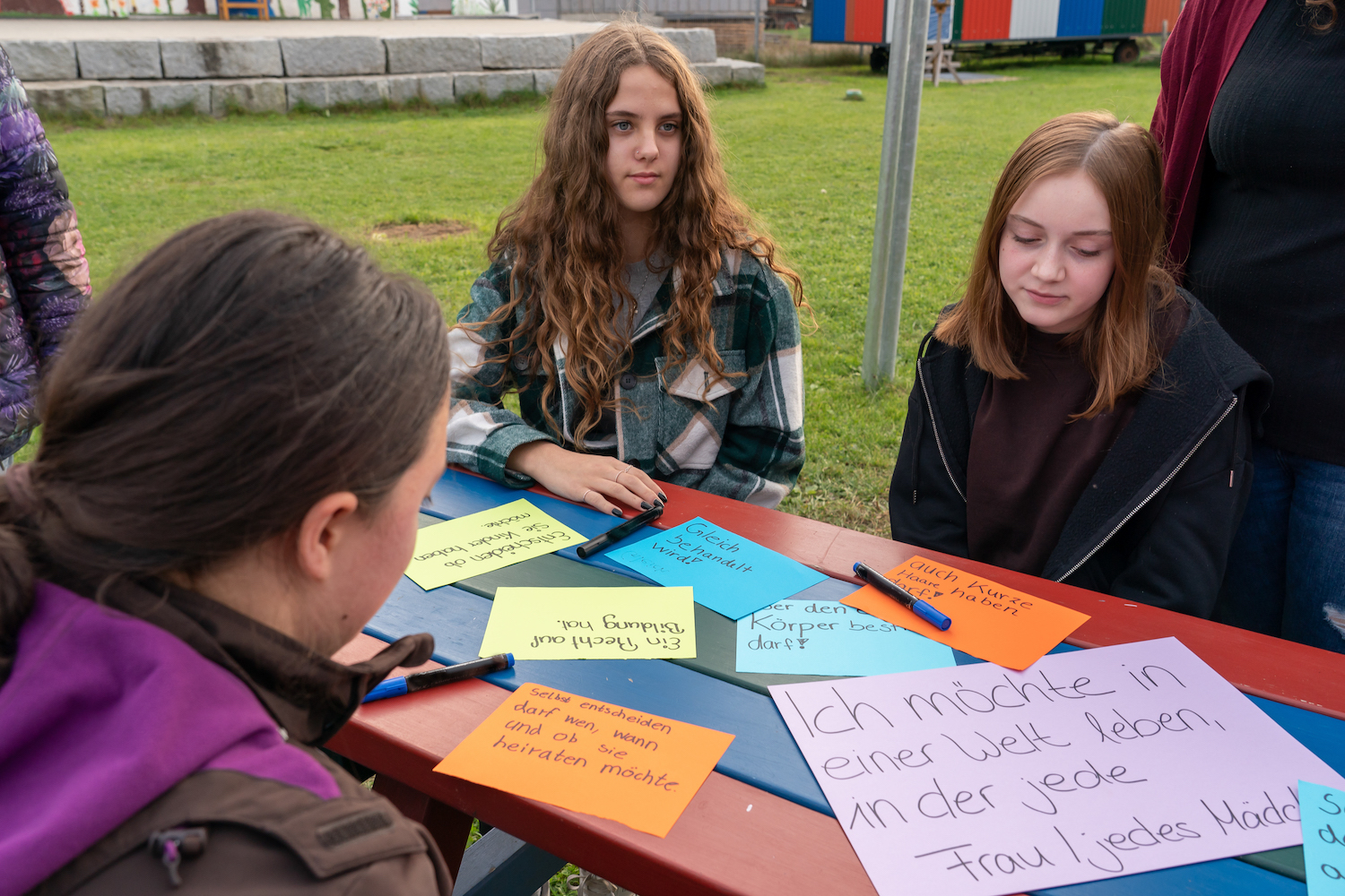 Drei Jugendliche sitzen an einem Tisch auf einer Wiese, auf dem Zettel mit verschiedenen Wünschen für Mädchen und Frauen auf der ganzen Welt