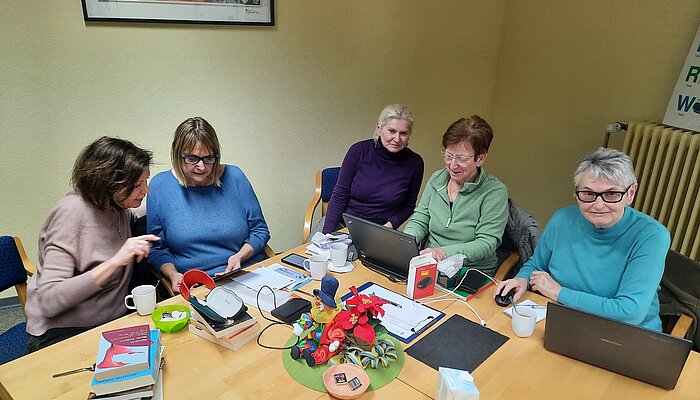 Fünf Frauen sitzen an einem Tisch und schauen auf Handybildschirme und Laptops