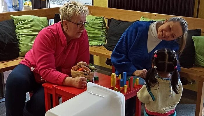 Zwei Frauen spielen mit einem Kleinkind an einem Tisch mit Bauklötzen