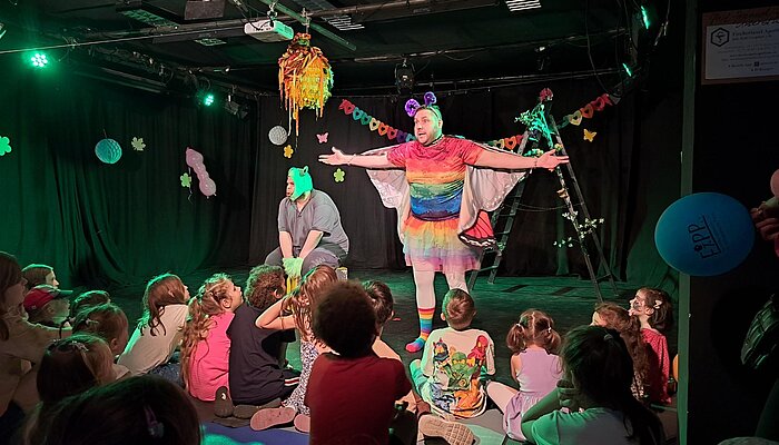 Das Bild zeigt eine Theateraufführung für Kinder. Ein Mann im Schmetterlingskostüm steht vor dem Publikum.