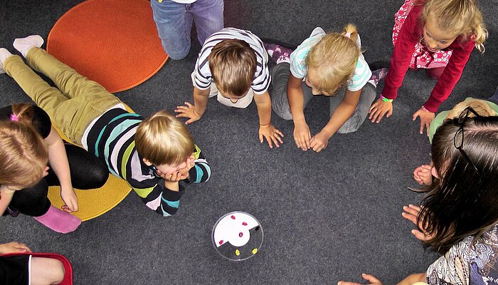 Acht Kinder und eine Frau hocken im Kreis auf dem Boden, in der Mitte steht eine Schale mit Fruchtgummi