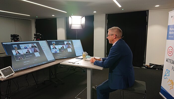 Dr. Winfried Kösters sitzt an Tisch mit Bildschirmen, auf denen Videokonferenzen  zu sehen sind