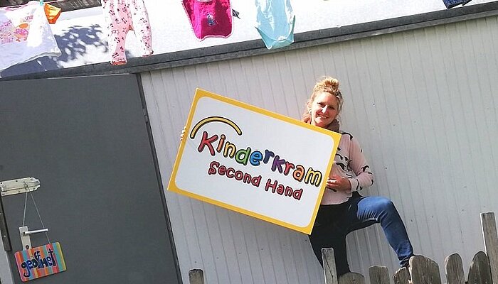 Frau steht vor einem Haus mit einem Schild in der Hand auf dem steht Kinderkrams Second Hand