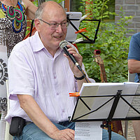 Ein älterer Mann sitzt draußen auf einem Stuhl und singt in ein Mirkofon. Vor ihm steht ein Notenständer. 