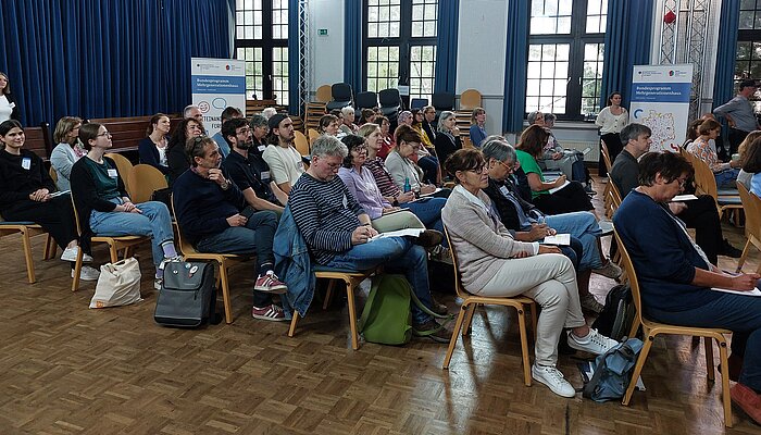 Mehrere Menschen sitzen in Stuhlreihen in einem großen Raum und folgen eine Rede