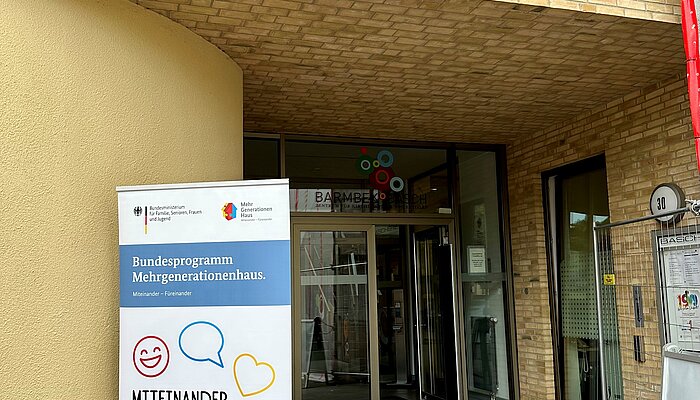 Eingang eines Gebäudes mit davorstehendem Roll-up mit der Aufschrift "Bundesprogramm Mehrgenerationenhaus. Miteinander – Füreinander"