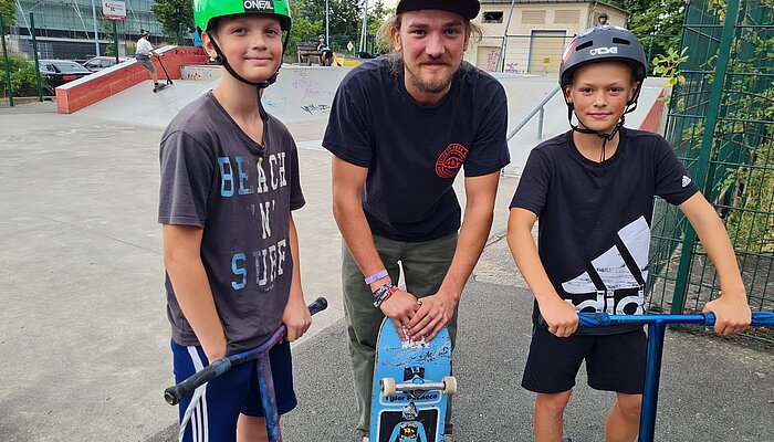 Drei Jungs tragen Helme, stehen auf ihren Rollern und lächeln.