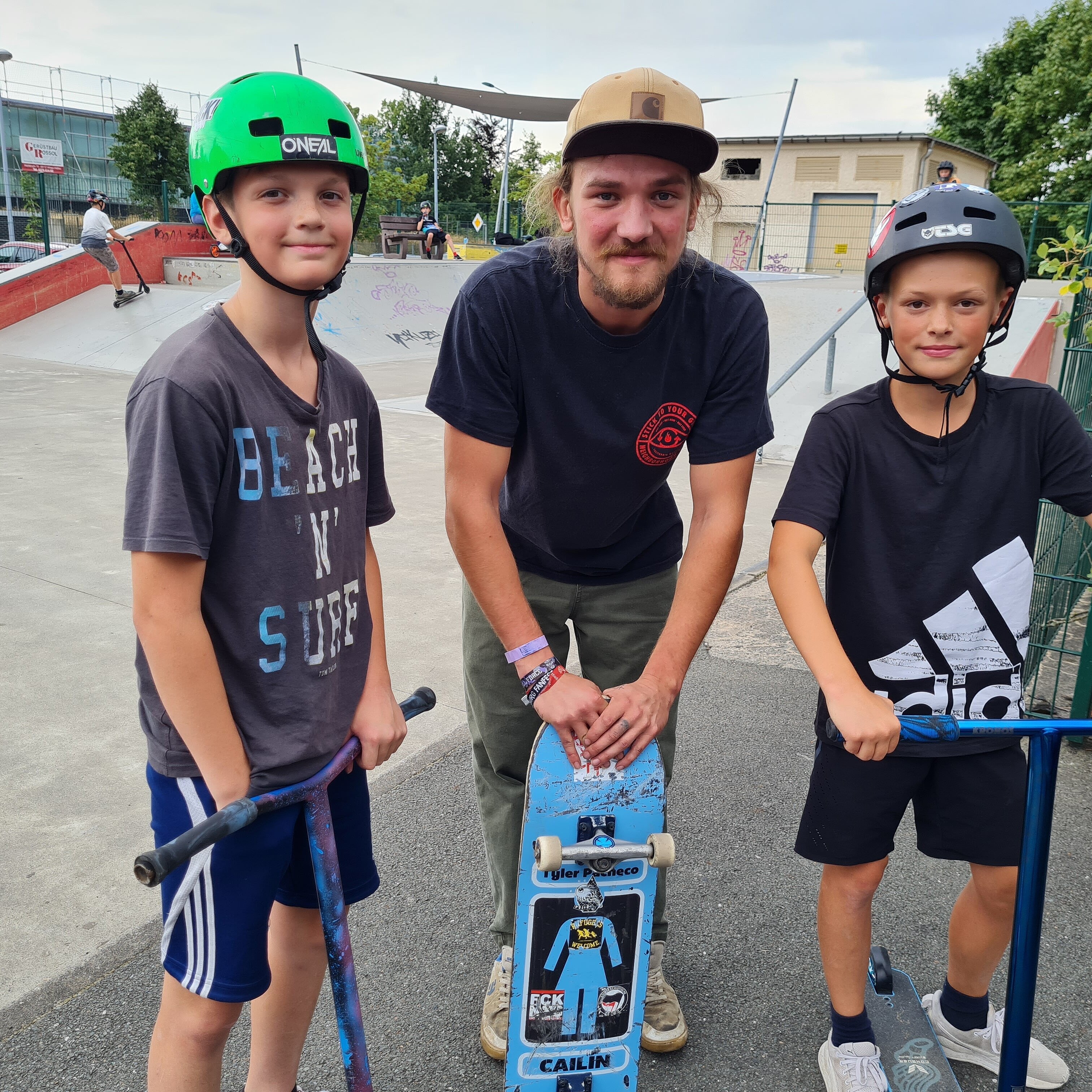 Drei Jungs tragen Helme, stehen auf ihren Rollern und lächeln.