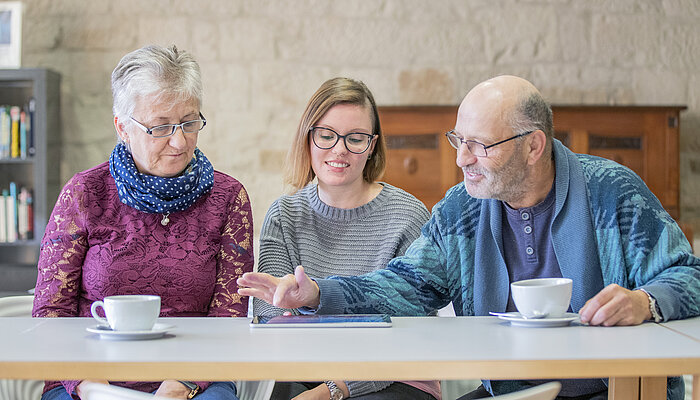 Seniorinnen und Senioren sitzen zusammen mit einer jungen Frau lächelnd an einem Tisch über ein Dokument gebeugt