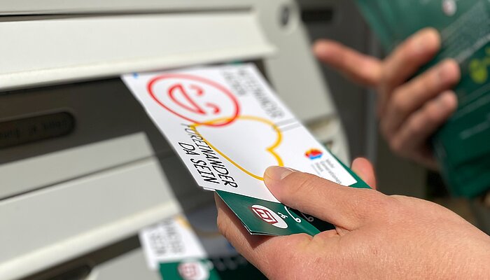 Eine Hand steckt eine Postkarte in einen Briefkasten.