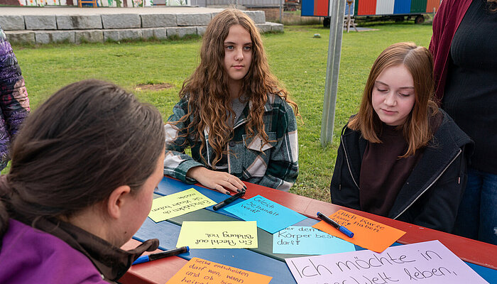 Drei Jugendliche sitzen an einem Tisch auf einer Wiese, auf dem Zettel mit verschiedenen Wünschen für Mädchen und Frauen auf der ganzen Welt