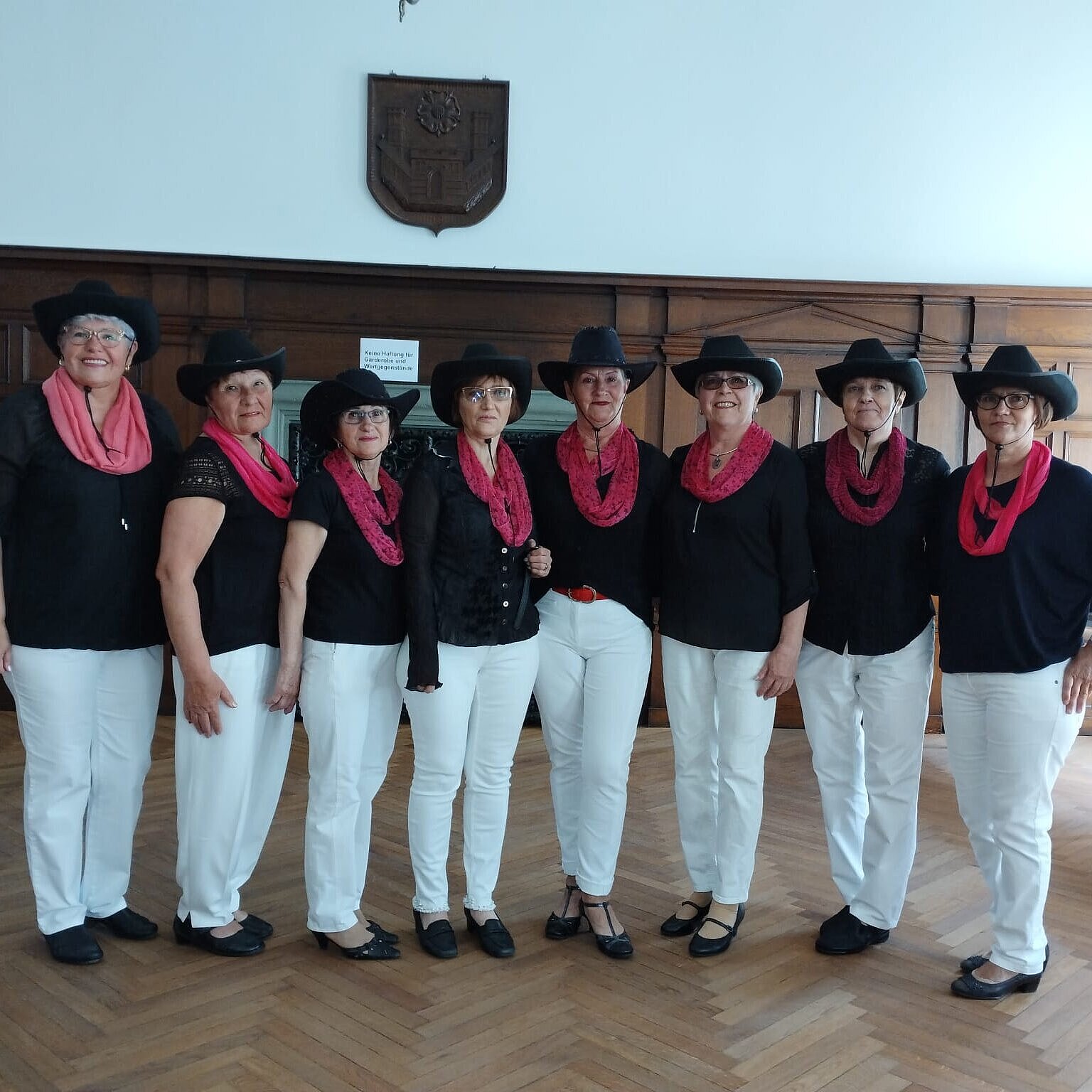 Acht Frauen mit Kleidung im Western-Stil stehen nebeneinander in einem holzverkleideten Saal