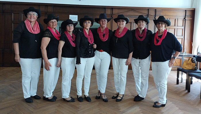 Acht Frauen mit Kleidung im Western-Stil stehen nebeneinander in einem holzverkleideten Saal